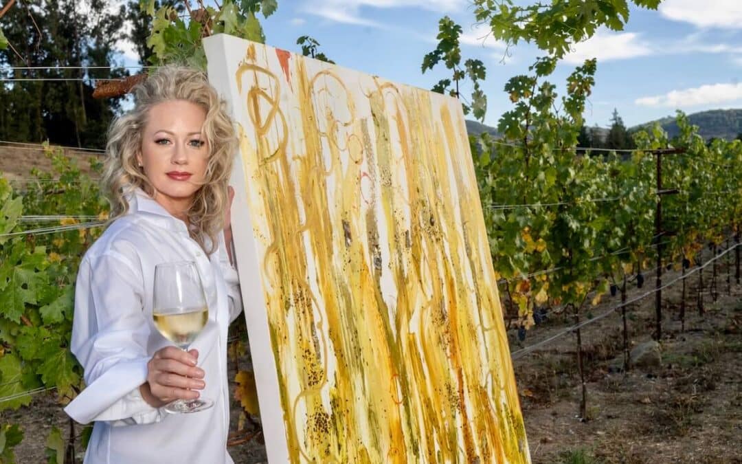 penelope moore holding glass of wine standing by paiting