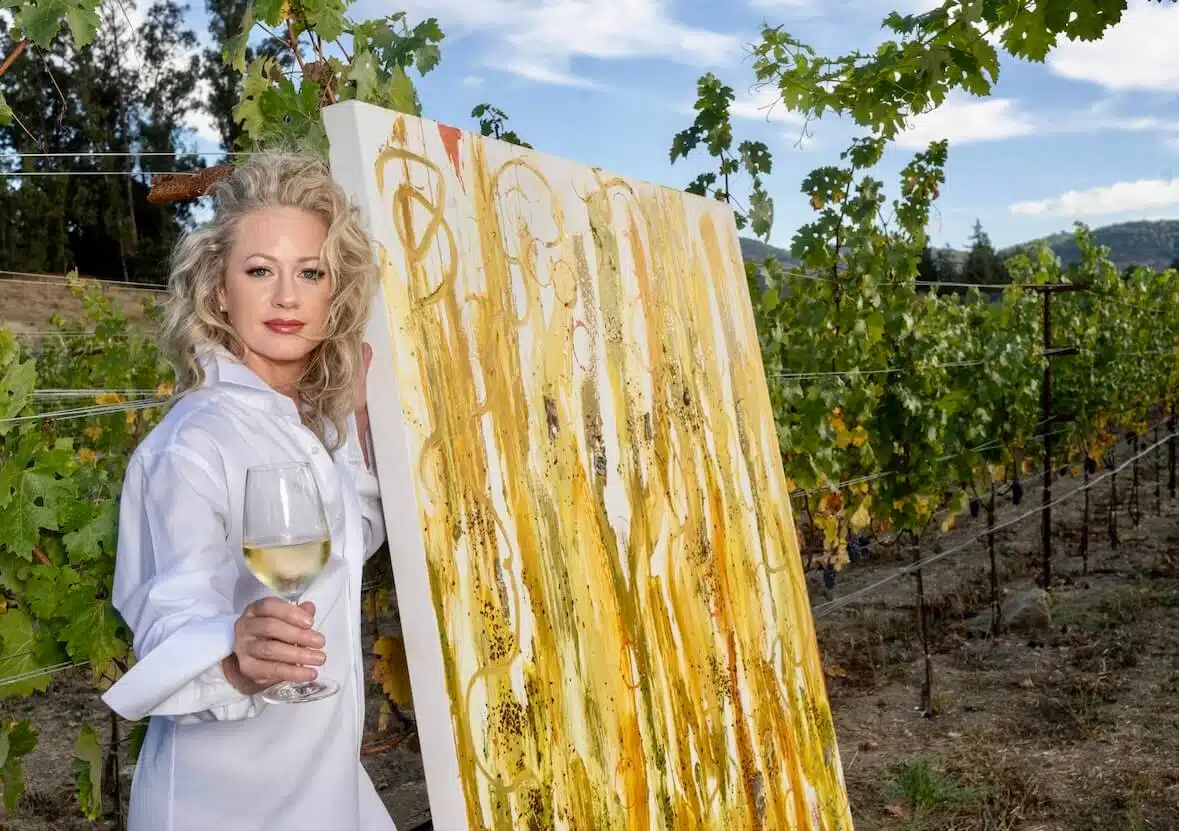 penelope moore holding glass of wine standing by paiting